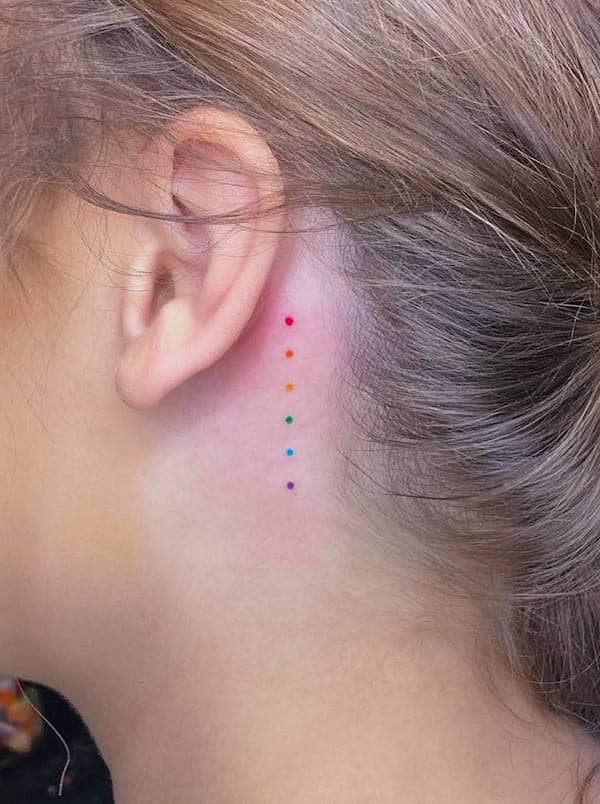 Small dots in rainbow colors behind the ear by @amandahowtattoo